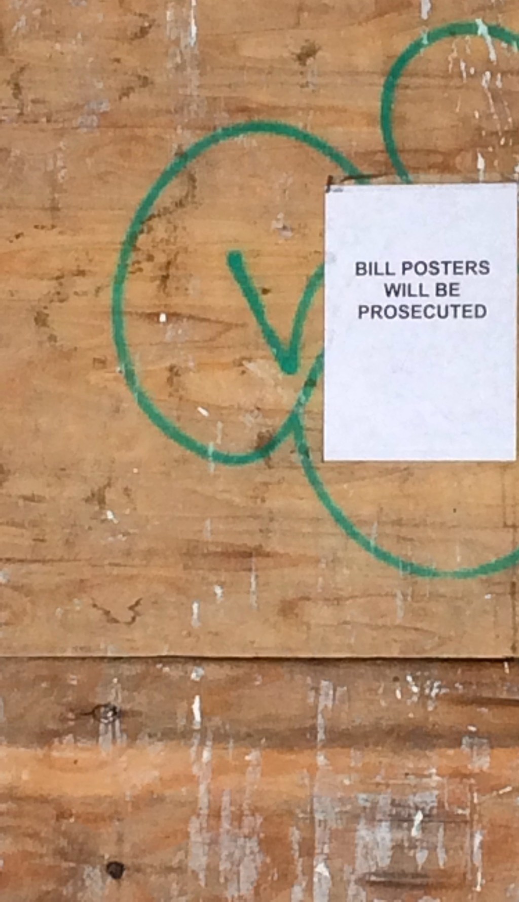 A notice on a plywood hoarding reading Bill posters will be prosecuted, beside green graffiti