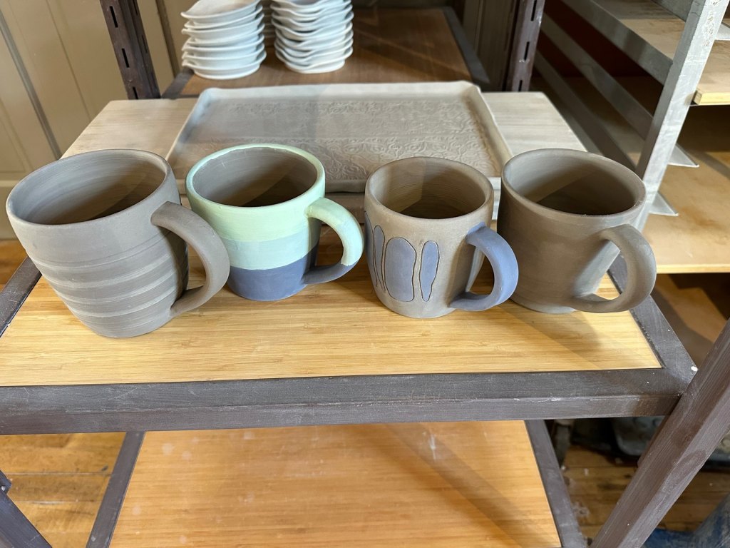 Four unfired mugs drying on a studio shelf