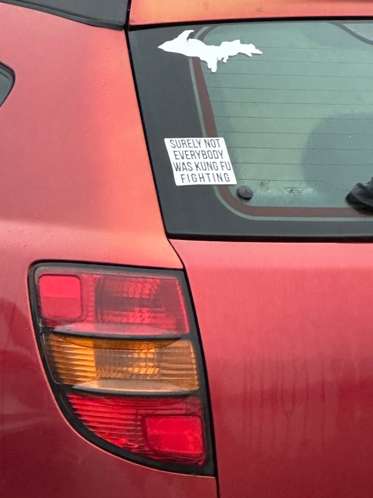 A bumper sticker on a red car reading Surely not everybody was kung fu fighting