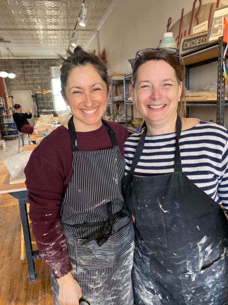 Robyn and Angela Patterson in clay-spattered aprons, smiling in the pottery studio